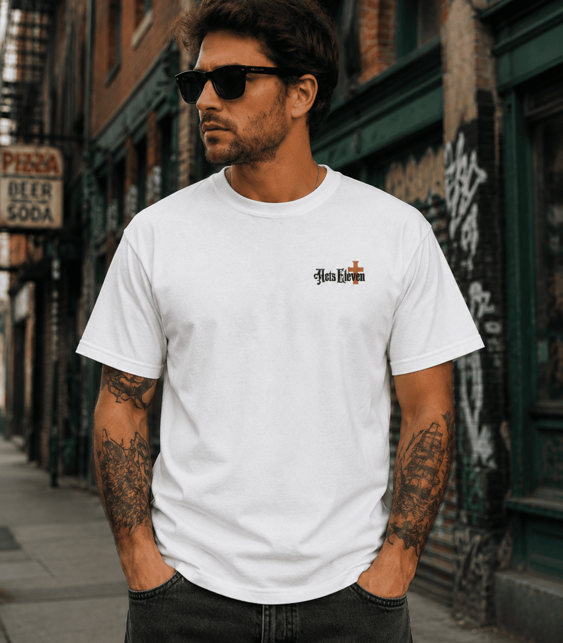 A man wearing a white short-sleeve t-shirt with a small gothic-style 'Heb Eschat' graphic on the left chest, standing on a city street.