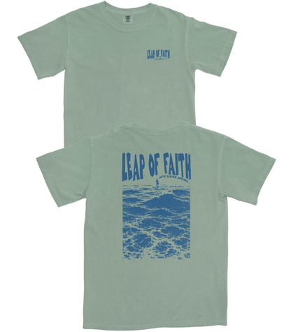 A sage green short-sleeve t-shirt shown from both the front and back. The front features a small blue 'Leap of Faith' logo on the left chest, while the back displays a large blue graphic of ocean waves with the text 'LEAP OF FAITH' arched over them.