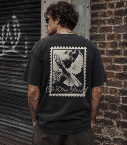 A man wearing a faded black t-shirt with a large white graphic print of a dove and the text 'A Olive Brand' on the back, standing in front of a brick wall and metal shutter.