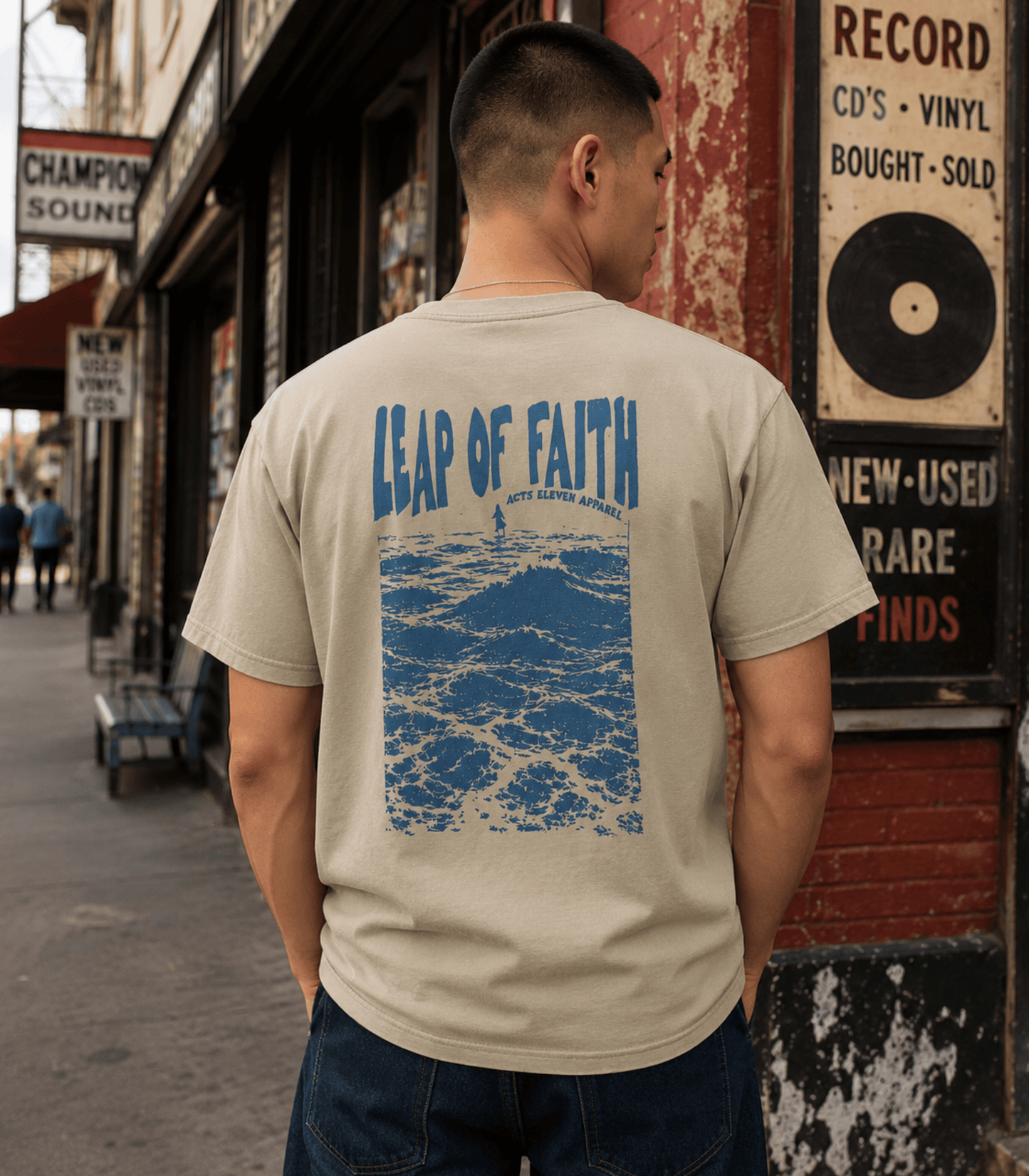 A man viewed from behind wearing a beige t-shirt with a blue graphic print that reads 'LEAP OF FAITH' above a blue ocean wave design. He is standing on a city street next to a brick wall with vintage record store signs.