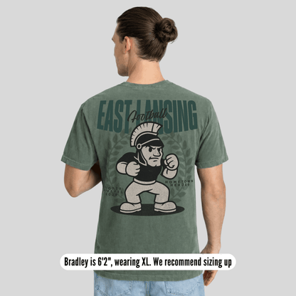 Person wearing a green t-shirt with 'East Lansing Football' and a cartoon character on the back, on a gray background.